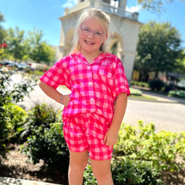 Young girl in a pink checkered outfit standing outdoors with a building and trees in the background.