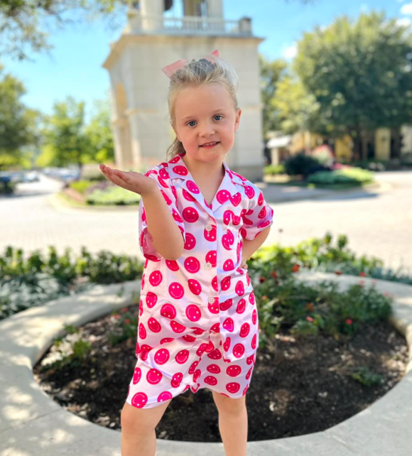 Young girl in a pink polka dot outfit standing outdoors with a building and trees in the background.